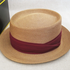 Pu&ograve; includere: Un cappello di paglia color beige con un'ampia tesa e un nastro bordeaux attorno alla corona.