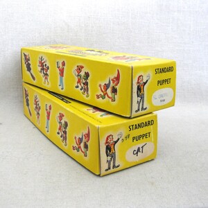May include: Two yellow rectangular boxes labeled "STANDARD PUPPET" with cartoon illustrations. One box is labeled "CAT" and the other "S. GRETTO". The boxes have a vintage aesthetic.