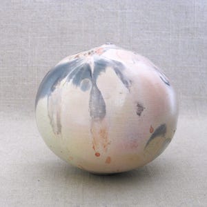 May include: A round ceramic vase with a speckled, organic design. The vase features a blend of colors, including cream, pale pink, and gray, with subtle brown accents. The overall shape is smooth and spherical, with a small opening at the top.