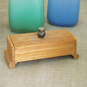 May include: A wooden box with a hinged lid and a small metal knob on top. The box is sitting on a beige fabric surface.