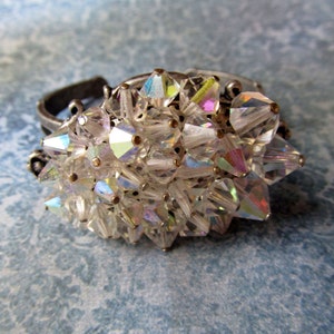 May include: A silver ring with a cluster of iridescent clear crystals. The crystals are arranged in a circular shape and are held together by a silver band.