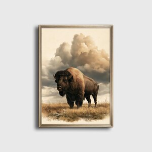 Può includere: Una stampa d'arte incorniciata raffigurante un grande bisonte marrone in piedi in un campo di erba secca e dorata. Lo sfondo mostra un cielo drammatico e nuvoloso con tenui toni beige e grigi. L'opera è racchiusa in una sottile cornice dorata metallica.