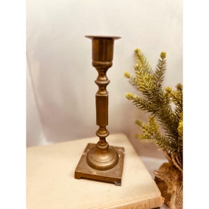 May include: A vintage brass candlestick holder with a square base and a detailed, ornate design. The candlestick has a cup-shaped top and a long, slender stem. The brass has a warm, aged patina. A small evergreen plant is in the background.