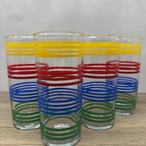 May include: A set of four clear glass tumblers, each featuring horizontal stripes in yellow, red, blue, and green. The cylindrical drinking glasses have a smooth finish and are arranged in a cluster, showcasing the colorful ring patterns.