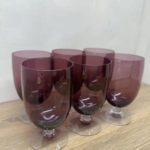 May include: A set of six purple glass goblets with clear glass stems and circular bases. Each goblet features a rounded bowl shape and a smooth, translucent finish, arranged in a cluster to display the deep violet color of the glass.