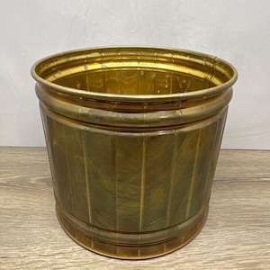 May include: A cylindrical brass wastebasket featuring a polished metallic finish with vertical ribbed detailing along the exterior body. The container has a wide open top with a reinforced rim and a smooth, circular base, showcasing a warm golden hue.