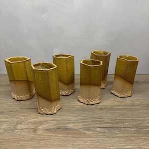 May include: A set of six hexagonal ceramic tumblers featuring a two-tone glaze. The upper portion is a speckled mustard yellow, while the lower section is a matte beige. Each tumbler has a scalloped, textured base edge and an open top.