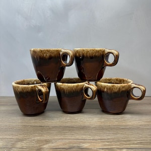 May include: A set of five ceramic cups arranged in a pyramid formation. Each cup features a deep brown base color that transitions into a lighter, creamy beige glaze around the rim, complete with a small, circular handle on the side.