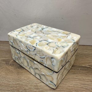 May include: A rectangular storage box featuring a mosaic exterior composed of iridescent, cream-colored shell fragments. The lid and sides are covered in smooth, polished pieces of shell with dark gray and white edges, creating a textured pattern.