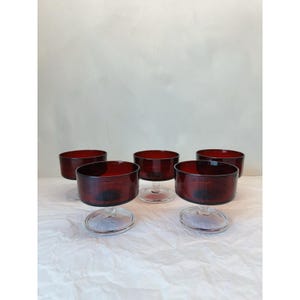 May include: A set of five deep red glass dessert bowls, each featuring a wide, circular basin and a short, clear glass pedestal base. The bowls are arranged in a cluster, showcasing their translucent ruby-colored glass and smooth, rounded rims.