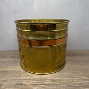May include: A cylindrical brass bucket featuring a polished finish and a prominent horizontal copper band encircling the middle section. The container has a wide open top, a smooth rounded rim, and a flat base, showcasing a metallic gold and copper color palette.
