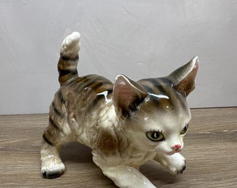 May include: A ceramic figurine of a tabby kitten in a pouncing stance. The kitten has brown and gray striped fur on its back and head, with a white chest, belly, and paws. It features green eyes, a pink nose, and a tail held upright in the air.