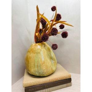 May include: A ceramic vase with a unique, organic shape, glazed in shades of yellow and green. The vase holds dried floral stems with burgundy ball-shaped blooms and golden-brown leaves. The vase rests on a stack of vintage books.