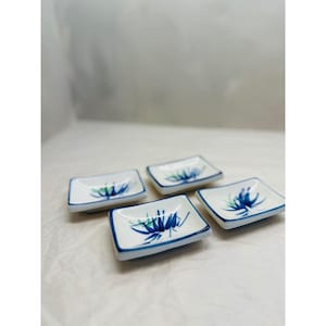 May include: Four small, square ceramic dishes. Each dish has a white base with a blue and green floral design and a blue border. The dishes are arranged on a white surface.