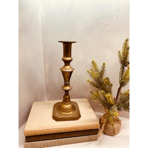 May include: A vintage brass candlestick holder with a tiered design. The candlestick has a wide, octagonal base and a tall, slender stem. The candlestick is sitting on top of a stack of books. A small evergreen plant is to the right.
