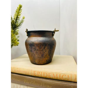 May include: A small, antique copper pot with a dark patina. The pot has a rounded body, a wide opening, and two small handles. It sits on top of a stack of old books. A sprig of greenery is visible in the background.