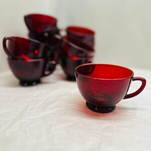 May include: A set of ruby red glass teacups with handles. The cups have a rounded bowl shape and sit on a small, circular base. The cups are arranged on a white surface.