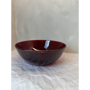 May include: A deep red glass bowl with a smooth, rounded rim and a subtle, swirling texture throughout the body. The bowl features a translucent quality that allows light to pass through, highlighting the rich, dark crimson color of the glass material.