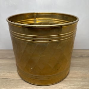 May include: A cylindrical brass planter featuring a textured diamond pattern across its exterior surface. The vessel has a smooth, polished rim and two horizontal embossed bands encircling the upper portion of the metallic body.