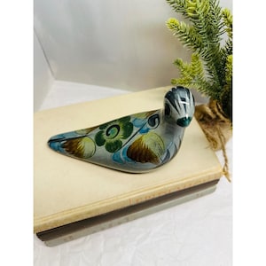 May include: A hand-painted ceramic bird figurine in shades of gray, blue, green, and brown. The bird is decorated with floral patterns and rests on a stack of books. A small potted plant is in the background.