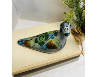 May include: A hand-painted ceramic bird figurine in shades of gray, blue, green, and brown. The bird is decorated with floral patterns and rests on a stack of books. A small potted plant is in the background.