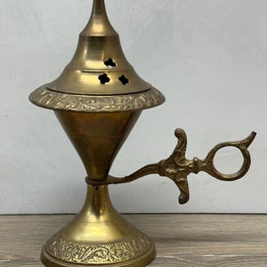 May include: A brass incense burner featuring a conical lid with small clover-shaped cutouts, a wide rim with embossed floral patterns, a tapered body, and a decorative handle with an ornate, scroll-like design attached to the base.