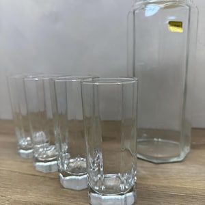 May include: A clear glass pitcher with a lid and four matching tall, hexagonal-shaped drinking glasses. The pitcher features a small gold-colored rectangular label on the upper side, and all items have a faceted, geometric design.
