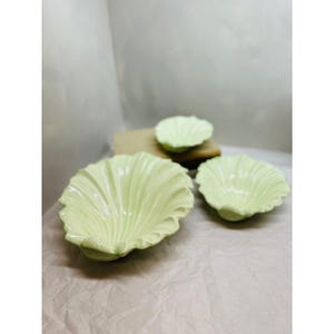 May include: Three light green ceramic bowls with a ruffled, leaf-like design. The bowls vary in size, with the largest in the foreground and two smaller ones in the background. The bowls are arranged on a light-colored surface.