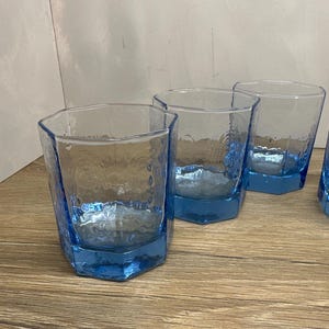 May include: A set of three light blue glass tumblers with a hexagonal shape and a textured, hammered finish on the exterior surface. Each glass features a thick, solid base and clear, translucent walls that catch the light, creating a distinct pattern.