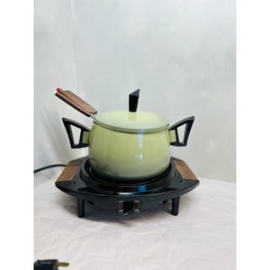 May include: A vintage fondue set with a light green pot, black handles, and a wooden base. The pot has a black lid and is set on a black electric warmer with a wooden trim. The set includes fondue forks with brown handles.