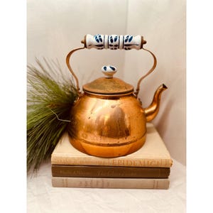 May include: A vintage copper tea kettle with a white and blue ceramic handle and lid knob. The kettle sits atop a stack of old books. The copper has a warm, aged patina. The handle is arched and made of copper.