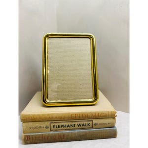 May include: A rectangular picture frame with a polished gold-tone metallic finish and rounded corners. The frame features a clear glass front and a neutral-colored backing insert, presented in a vertical orientation for displaying photographs.