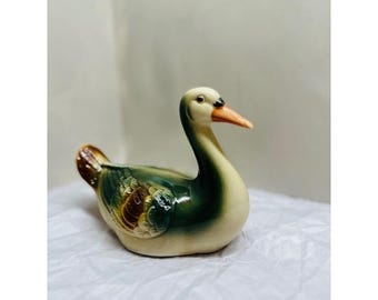 May include: A ceramic figurine of a duck with a glossy finish. The duck features a cream-colored head and neck, a bright orange beak, and a body with blended shades of dark green and brown feathers. The duck is depicted in a seated position.