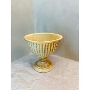 May include: A cream-colored pedestal bowl featuring a wide, flared rim and a vertical ribbed texture. The exterior surface displays a distressed, brownish-gold finish applied over the cream base, highlighting the raised vertical ridges.