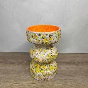 May include: A cylindrical ceramic vase with a stacked, three-tiered design. The exterior features a textured, speckled finish in shades of yellow, black, and white. The interior of the vase is a solid, bright orange with a speckled texture.