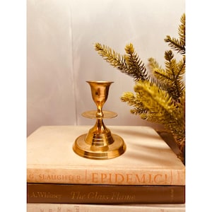 May include: A brass candlestick holder with a round base, a small circular plate, and a cup-shaped top. The candlestick is placed on top of a stack of old books. A sprig of yellow-green foliage is in the background.