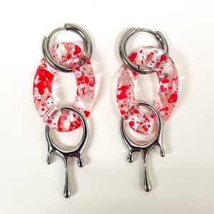 May include: A pair of silver hoop earrings with a dangling drop design. The hoops are adorned with clear acrylic charms that have a pink splatter pattern.