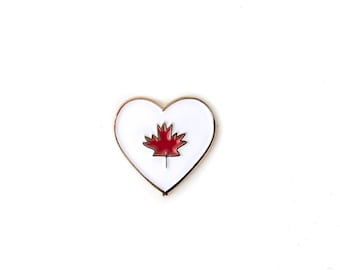 May include: A white heart-shaped pin with a red maple leaf in the center. The pin has a gold outline.