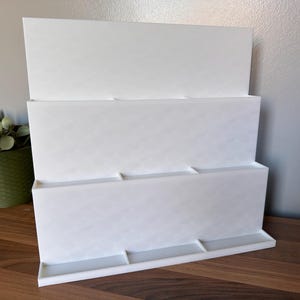 May include: A white, three-tiered display stand with a rectangular base and three shelves. The stand is made of a solid, opaque material and is designed to hold items. The stand is on a wooden surface.