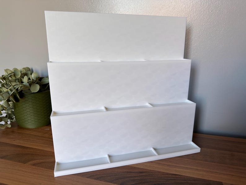 May include: A white, three-tiered display stand with a rectangular base and three shelves. The stand is made of a solid, opaque material and is designed to hold items. The stand is on a wooden surface.