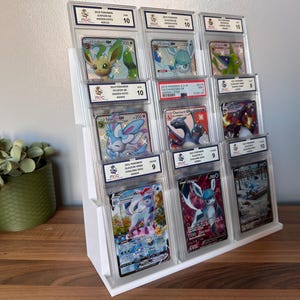 May include: A white tiered display stand holding multiple graded Pok&eacute;mon trading cards. The cards feature various Pok&eacute;mon characters, including Sylveon, Glaceon, and Charizard. Each card is encased in a protective holder with a grade label.