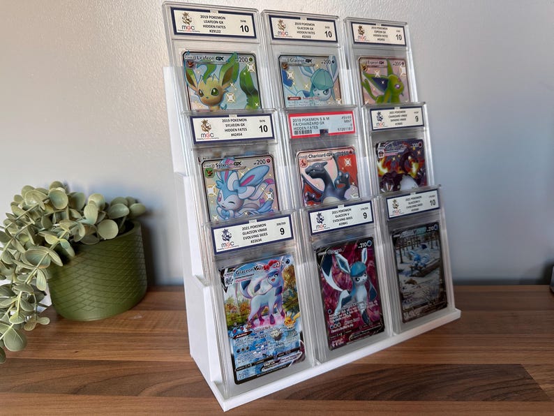 May include: A white tiered display stand holding multiple graded Pok&eacute;mon trading cards. The cards feature various Pok&eacute;mon characters, including Sylveon, Glaceon, and Charizard. Each card is encased in a protective holder with a grade label.