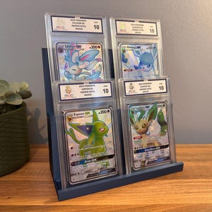 May include: A blue display stand holding four graded Pok&eacute;mon cards. The cards feature various Pok&eacute;mon characters, including Sylveon, Glaceon, Espeon, and Leafeon. Each card is encased in a protective holder with a grade of 10.