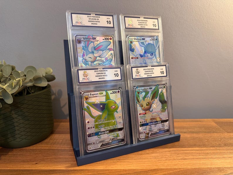May include: A blue display stand holding four graded Pok&eacute;mon cards. The cards feature various Pok&eacute;mon characters, including Sylveon, Glaceon, Espeon, and Leafeon. Each card is encased in a protective holder with a grade of 10.