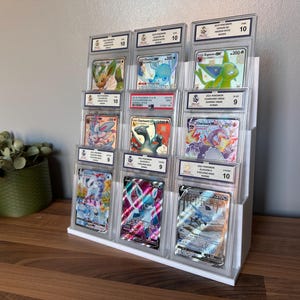 May include: A clear acrylic display stand holding graded Pok&eacute;mon trading cards. The stand is on a wooden surface. The cards feature various Pok&eacute;mon characters and are encased in protective holders with grading information.
