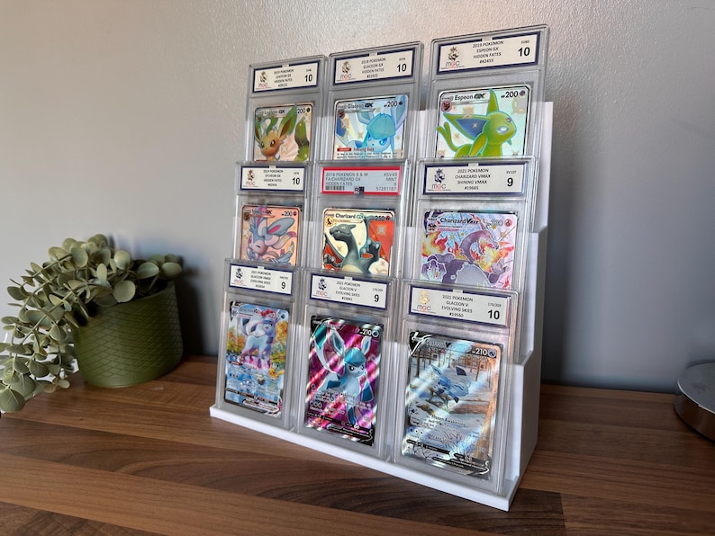May include: A clear acrylic display stand holding graded Pok&eacute;mon trading cards. The stand is on a wooden surface. The cards feature various Pok&eacute;mon characters and are encased in protective holders with grading information.