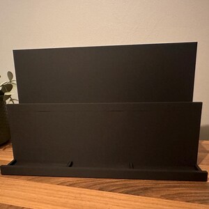 May include: A black desktop organizer with a tiered design. The organizer has a tray at the bottom and a back panel with two sections. The item is made of a matte material and sits on a wooden surface.