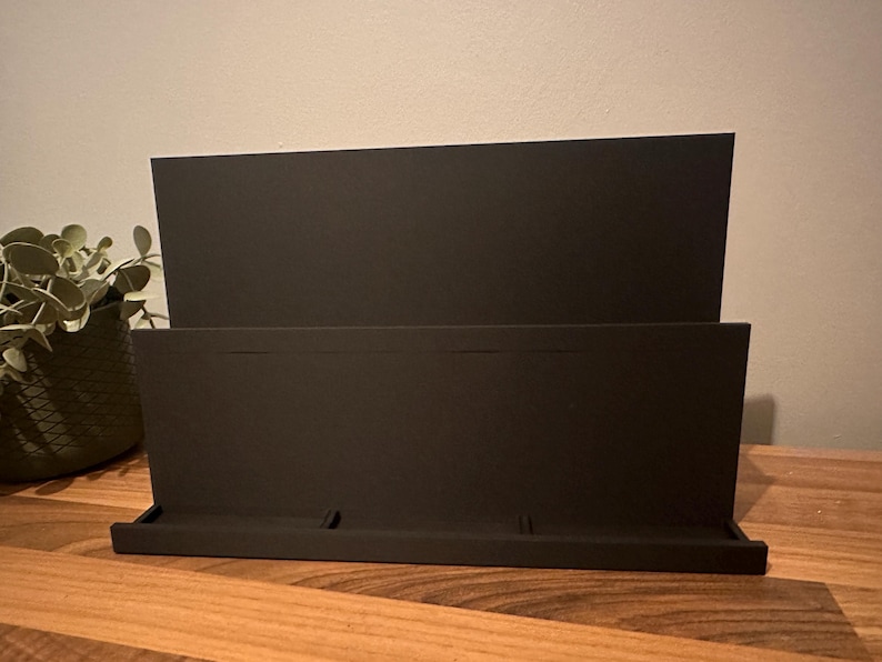 May include: A black desktop organizer with a tiered design. The organizer has a tray at the bottom and a back panel with two sections. The item is made of a matte material and sits on a wooden surface.