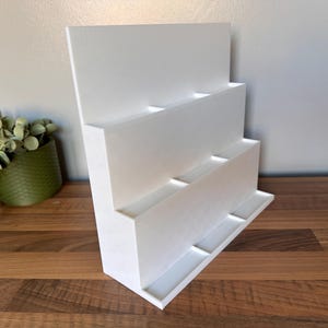 May include: A white, tiered display stand with multiple rectangular compartments. The stand is angled and sits on a wooden surface. A green potted plant is visible to the left.