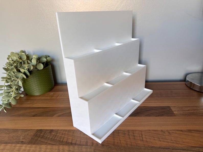 May include: A white, tiered display stand with multiple rectangular compartments. The stand is angled and sits on a wooden surface. A green potted plant is visible to the left.
